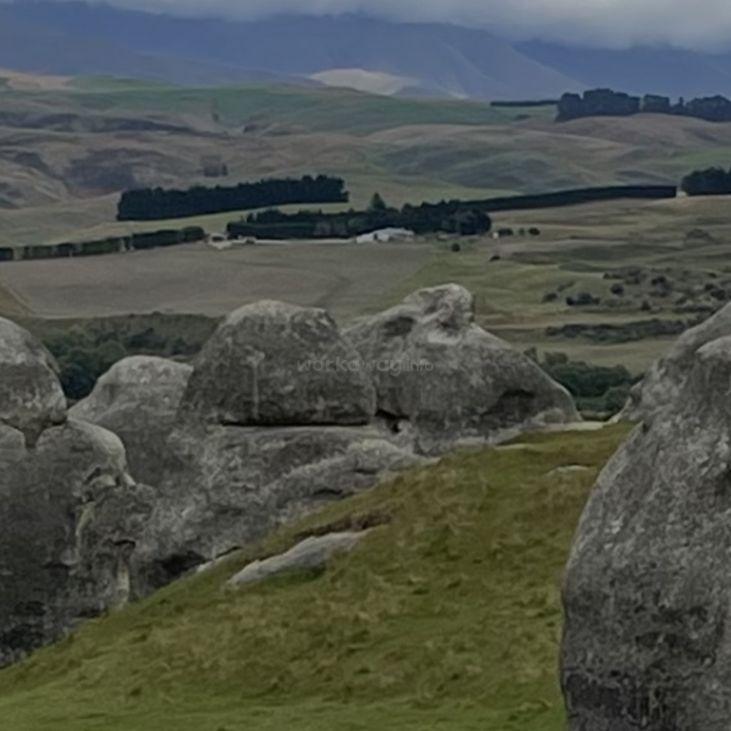 De imposantes formations rocheuses grises dominent le premier plan, entourées de collines verdoyantes. À l'arrière-plan, des champs, des rangées d'arbres sombres et des montagnes brumeuses composent un paysage serein.