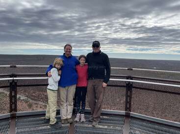 Eine vierköpfige Familie steht zusammen auf einer Aussichtsplattform, umgeben von einer weitläufigen Landschaft unter einem bewölkten Himmel, und genießt gemeinsam ein malerisches Outdoor-Abenteuer.