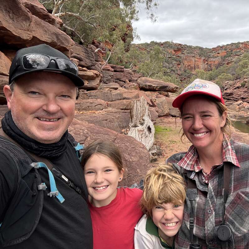 Uma família feliz de quatro pessoas posa junta ao ar livre, cercada por terrenos rochosos e árvores. Eles estão sorrindo, vestidos de forma casual e curtindo uma aventura na natureza juntos.
