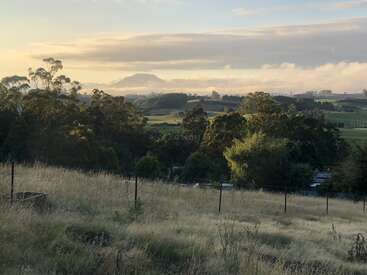 A peaceful rural landscape at sunrise, featuring grassy fields, scattered trees, distant mountains, a misty horizon, and a fence in the foreground. Tranquil countryside scene.