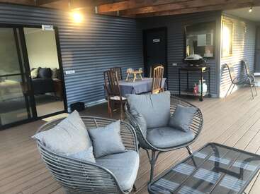 A cozy outdoor patio with cushioned chairs, a glass-top table, wooden dining set, barbecue, fire extinguisher, and sliding doors leading into a bedroom. Warm lighting.