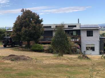 A modern two-story house with solar panels sits in a rural area. A black SUV is parked nearby. Trees and dry grass surround the property.