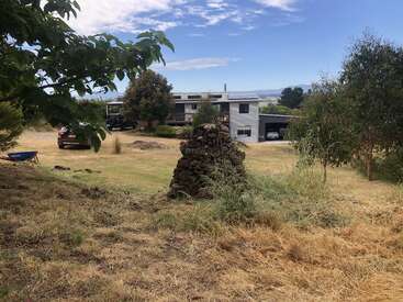 A modern house sits in a rural landscape with dry grass, trees, parked cars, and a rock pile in the foreground under a partly cloudy sky.