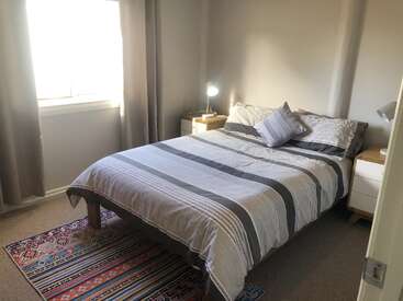 This cozy bedroom features a double bed with striped bedding, two bedside tables with lamps, a colorful patterned rug, and sunlight streaming through a curtained window.