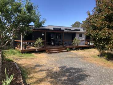 A modern single-story house with a large wooden porch surrounded by plants and trees. There is a gravel driveway and patches of grass in the yard.