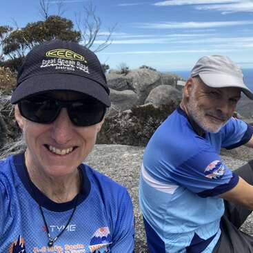 Two people sit on rocky terrain, smiling in matching blue shirts and hats. The background includes trees, large boulders, and a blue sky with clouds.