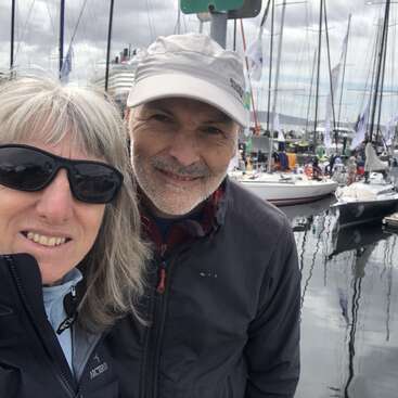 A smiling couple poses for a selfie at a marina with sailboats and people in the background. They appear happy, dressed warmly under a cloudy sky.