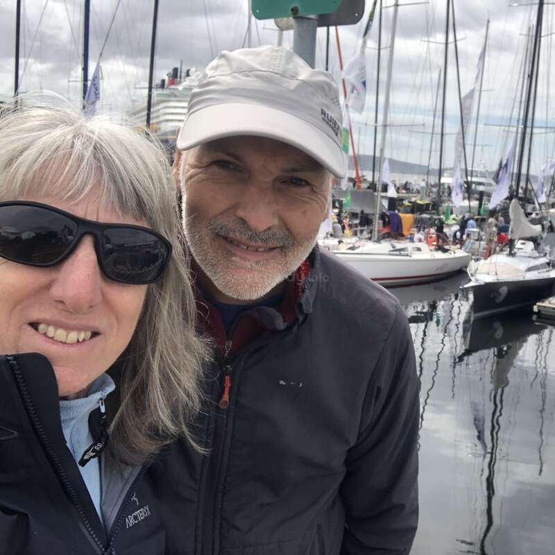 Un couple souriant pose pour un selfie dans une marina avec des voiliers et des gens en arrière-plan. Ils semblent heureux, vêtus chaudement sous un ciel nuageux.