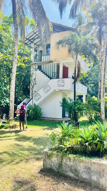 A two-story white house with outdoor stairs, surrounded by lush green trees and plants. A person is gardening or mowing the lawn in sunlight.