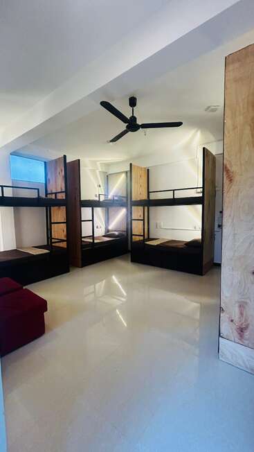 The image shows a modern dorm room with three sets of wood-framed bunk beds, a ceiling fan, white floors, minimal decor, and a maroon couch.