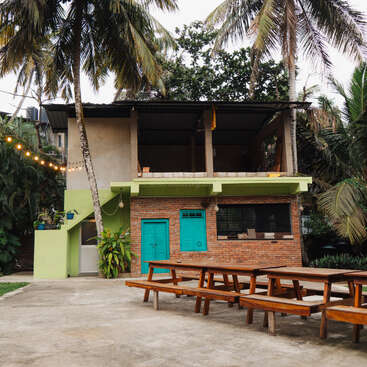 Ein kleines Backsteinhaus mit türkisfarbenen Türen steht zwischen Palmen. Draußen gibt's Picknicktische aus Holz, und Lichterketten sorgen für eine gemütliche, einladende tropische Atmosphäre.