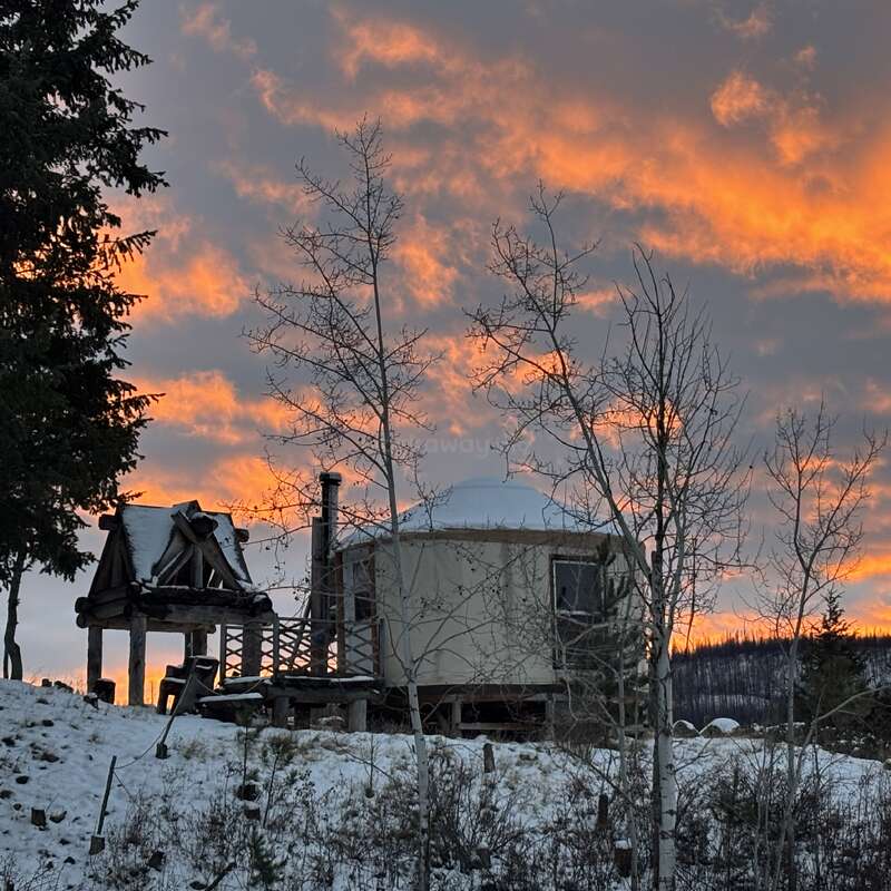 Eine gemütliche Hütte steht an einem verschneiten Hang, umgeben von kahlen Bäumen. Leuchtend orangefarbene Wolken erhellen den Abendhimmel und schaffen eine friedliche und einladende Winterkulisse.