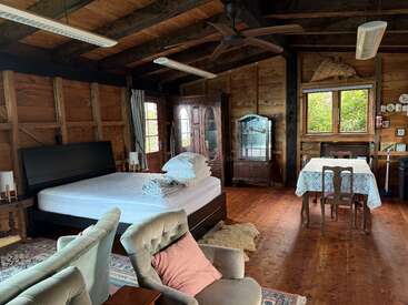 Cette chambre rustique et confortable comprend un lit, des fauteuils, des meubles vintage en bois, une table à manger avec une nappe en dentelle, des murs en bois et de grandes fenêtres qui laissent entrer la lumière naturelle.