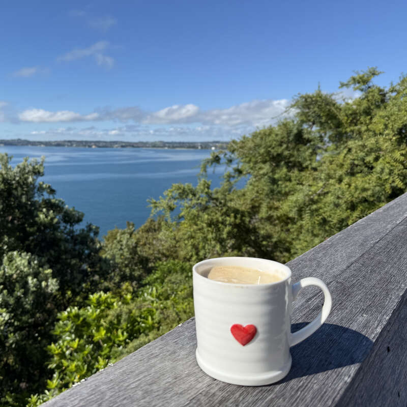 Una taza blanca con un corazón rojo se asienta sobre una barandilla de madera, con vistas a un sereno lago azul, rodeado de verdes árboles bajo un brillante cielo soleado.