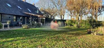Ein weitläufiger Garten mit grünem Rasen, ein paar abgefallenen Blättern und Bäumen. Eine Person fährt auf einem runden Gerät, das Nebel erzeugt. Man sieht ein Holzhaus mit Gartenmöbeln.