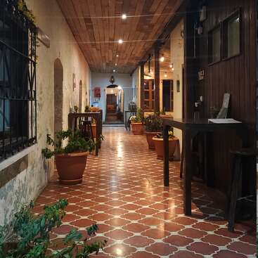 Un couloir confortable avec un sol carrelé, un plafond en bois et un éclairage chaleureux. Des plantes en pot et des tables bordent l'allée, créant une atmosphère accueillante dans un patio intérieur.