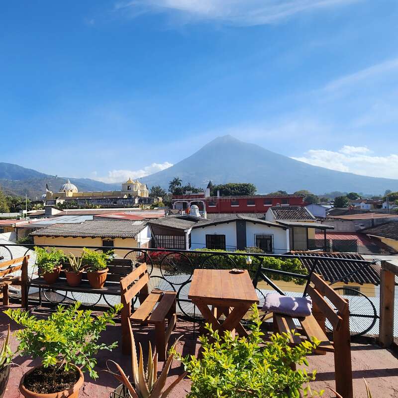 Um terraço ensolarado na cobertura, com mesas de madeira, vasos de plantas e bancos, tem vista para os edifícios coloridos da cidade e um vulcão incrível sob um céu azul claro. Ambiente tranquilo.