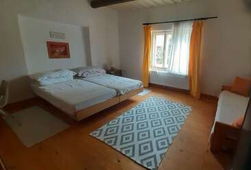 Se trata de un dormitorio acogedor y sencillo con dos camas individuales, un pequeño sofá, suelo de madera, alfombra estampada, cortinas amarillas, paredes blancas y una ventana iluminada por el sol.