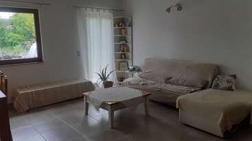 Un salón luminoso y acogedor con muebles de color beige, una mesa de centro, una maceta, una estantería, un gran ventanal, cortinas transparentes y baldosas neutras. Ambiente tranquilo.