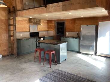 A imagem mostra uma cozinha moderna com painéis de madeira clara, aparelhos de aço inoxidável e armários cinza, com uma ilha e banquetas e uma área de loft acima.