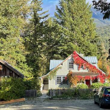 Das Bild zeigt ein charmantes zweistöckiges Haus mit einer hellen Fassade, umgeben von Bäumen und einer leuchtend roten Weinrebe, eingebettet in eine ruhige Berglandschaft.
