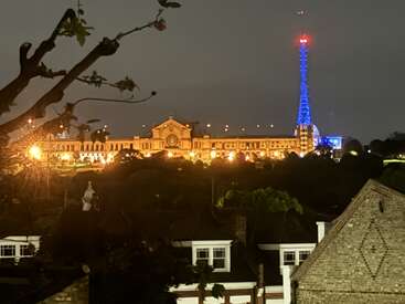 Ein großes, beleuchtetes Gebäude steht nachts auf einem Hügel, mit einem markanten, blau beleuchteten Turm in der Nähe. Im Vordergrund sind Baumzweige und Dächer unter dem dunklen Himmel zu sehen.
