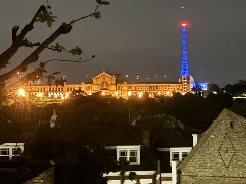 Ein großes, beleuchtetes Gebäude steht nachts auf einem Hügel, mit einem markanten, blau beleuchteten Turm in der Nähe. Im Vordergrund sind Baumzweige und Dächer unter dem dunklen Himmel zu sehen.