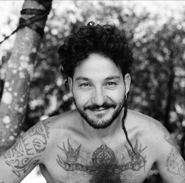 Ein lächelnder Typ mit lockigem Haar, Bart und Tattoos auf Brust und Armen posiert ohne Shirt draußen. Im Hintergrund steht ein Baum.