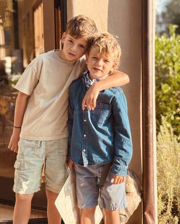 Two young boys stand closely together outdoors, one with his arm around the other. They both smile warmly, dressed casually in summer clothes, sunlight illuminating them.