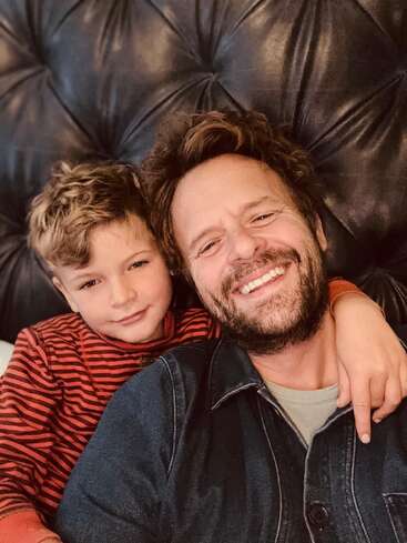 A man and a young boy are smiling and taking a selfie together. The boy has his arm around the man’s shoulder. They look happy.