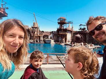 A family of four poses for a selfie at an outdoor theme park attraction featuring a large water set and industrial, post-apocalyptic structures under a blue sky.