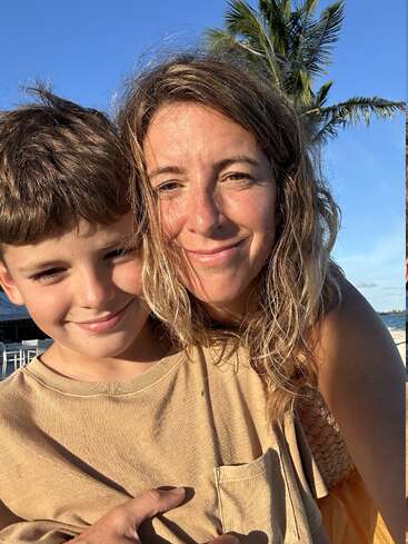 A woman and young boy smile for a close-up selfie on a sunny beach. Palm tree, blue sky, and ocean create a warm, happy vacation vibe.