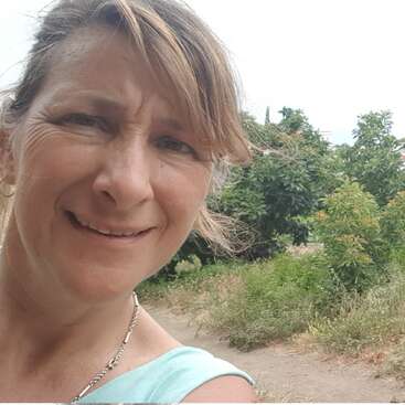 Uma mulher sorri para uma selfie em close-up ao ar livre. Atrás dela, árvores e plantas verdes exuberantes criam um fundo natural e tranquilo em um dia nublado.