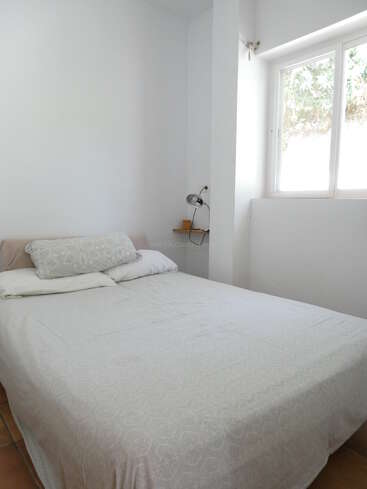 Esta imagem mostra um quarto pequeno e minimalista com uma cama de casal bem arrumada, roupa de cama branca, uma janela grande, luz natural, paredes brancas e um abajur.
