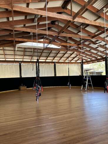 La imagen muestra una sala abierta con suelo de madera y vigas a la vista, en la que cuelgan varias cuerdas y una escalera, lo que sugiere un gimnasio o espacio para hacer ejercicio.