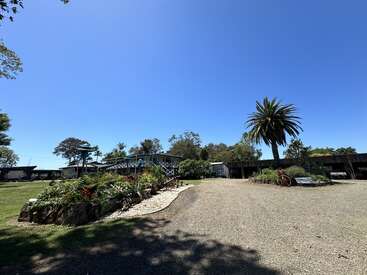 La imagen representa un paisaje sereno con una gran casa, un camino de grava y una palmera, todo ello bajo un cielo azul despejado.