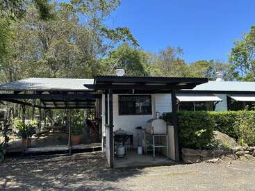 La imagen muestra un pequeño edificio blanco con un porche cubierto y una zona de cocina al aire libre, rodeado de árboles y un cielo azul despejado.