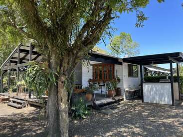 La imagen representa una encantadora casa blanca con un porche cubierto, rodeada de frondosos árboles y plantas, que desprenden una sensación de serenidad y belleza natural.