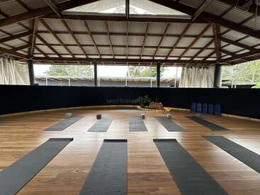 La imagen muestra una amplia sala con suelo de madera, en la que hay esterillas de yoga y bloques dispuestos en hileras, con una pared negra y un techo con vigas a la vista.