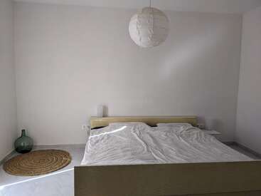 La imagen muestra un dormitorio sereno con una cama, paredes blancas y una alfombra redonda, con un gran farolillo de papel blanco suspendido sobre la cama.