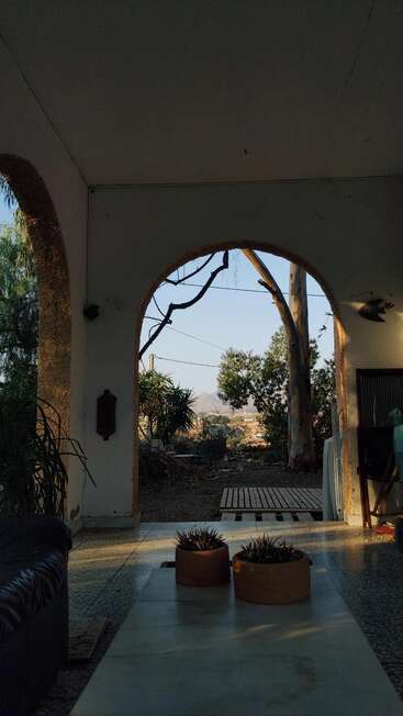 La imagen muestra un espacio exterior sereno con una puerta arqueada, un banco, plantas en macetas y una vista panorámica de árboles y montañas a lo lejos.