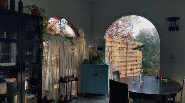 La imagen muestra una habitación con una mesa y sillas, una estantería, una lámpara y una ventana. La habitación está bien iluminada, con una combinación de colores neutros y una sensación de calma.
