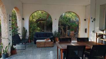 La imagen muestra un espacio exterior sereno con una mesa y sillas, rodeado de exuberante vegetación, que evoca una sensación de tranquilidad y relajación. El ambiente es apacible y acogedor.