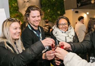 The image shows three people toasting with small glasses of red liquid, wearing lanyards and standing in front of a green wall with a large stone face.