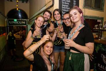 The image shows six people posing for a photo, each holding a drink, with a blackboard displaying alcohol prices in the background, likely at a bar or pub.