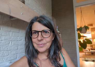 Una mujer con largas canas y gafas sonríe a la cámara. Está en un interior, con plantas, luz solar y una decoración acogedora visibles al fondo.