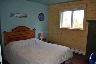 Cette chambre à coucher confortable se compose d'un lit en bois, de murs bleus et lambrissés, d'une grande fenêtre, d'un mobilier simple, d'une horloge murale et d'une baleine décorée au-dessus du lit.