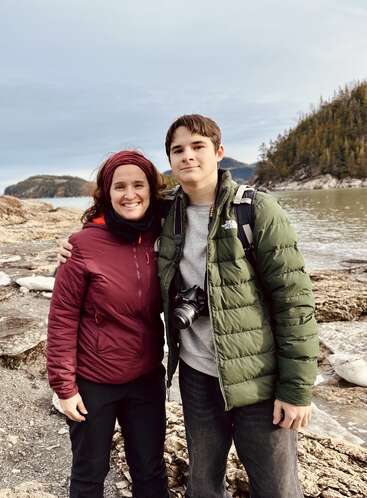 Une femme souriante et un jeune homme se tiennent à l'extérieur, près d'un rivage rocheux, vêtus de vestes. Il a un appareil photo et un sac à dos. Derrière eux, des collines boisées et de l'eau.