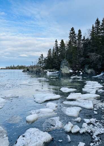 De gros morceaux de glace fondante flottent sur un lac calme et partiellement gelé. Des arbres à feuilles persistantes bordent le rivage rocheux sous un ciel bleu vif, partiellement nuageux.
