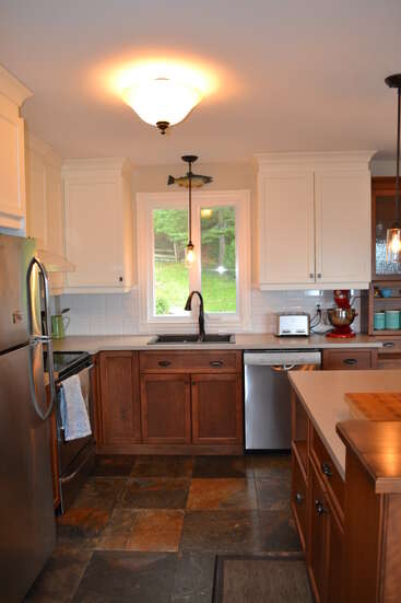 Cette cuisine se caractérise par des placards inférieurs en bois, des placards supérieurs blancs, des appareils électroménagers en acier inoxydable, une fenêtre au-dessus de l'évier, un éclairage suspendu, des carreaux de sol en ardoise et une atmosphère chaleureuse.